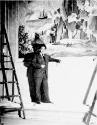 caption: N. C. Wyeth posing in front of mural during installation at the Penn Mutual Insurance …